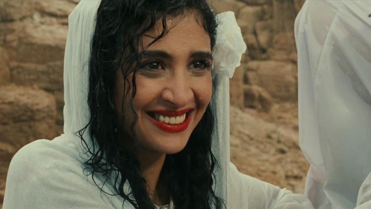 THE OTHER, 1999, woman wearing white dress with hood smiling and seated on rocky surface