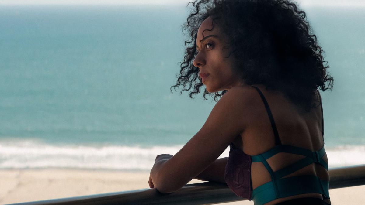 MISTY GREEN, 2026, woman in bathing suit leaning over ledge of balcony with ocean and beach in background