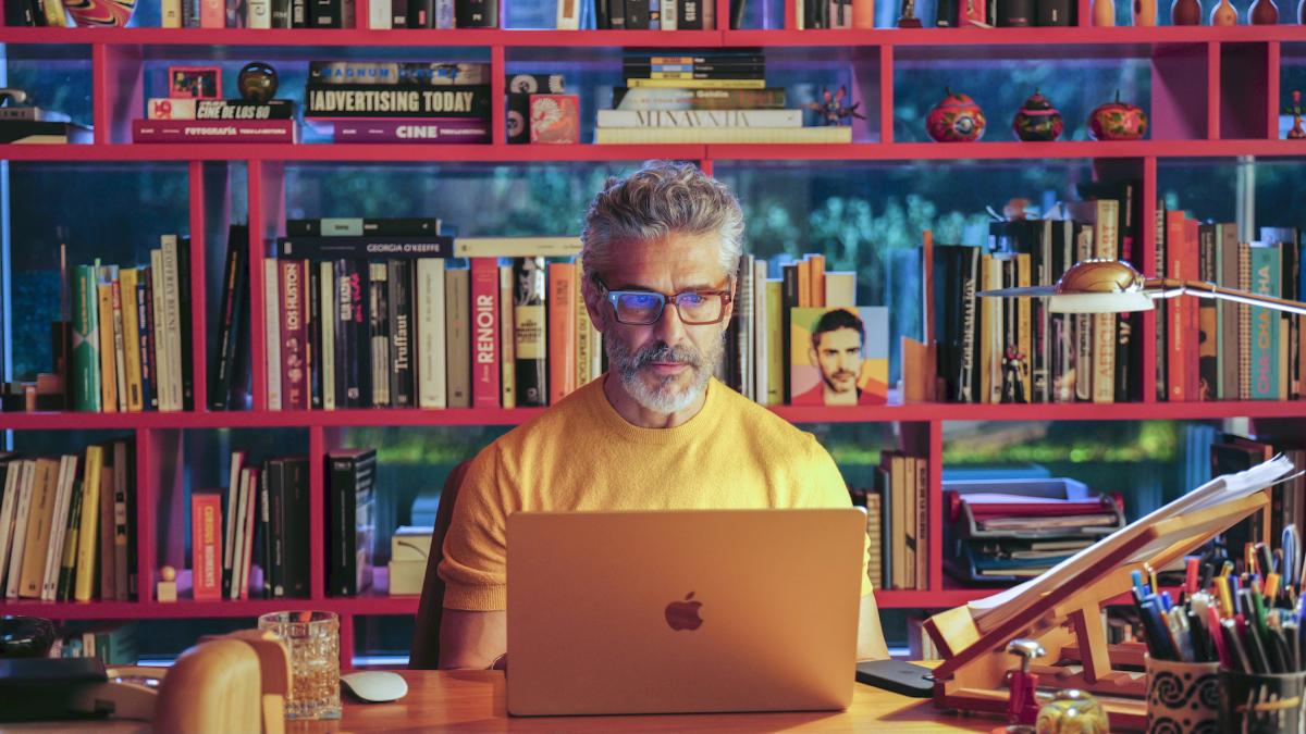 BITTER CHRISTMAS, 2026, man in yellow sweater wearing glasses and typing on laptop in front of large bookcase