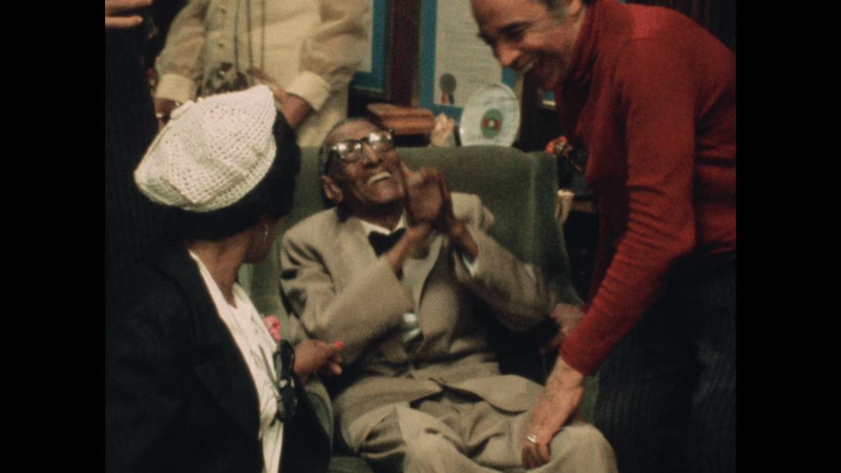 ONCE UPON IN HARLEM, 2026, people gathered in room leaning around and smiling person in chair