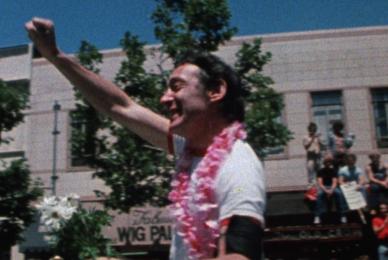 man in white shirt and lei holding hand up in fist in street parade