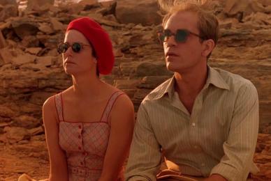 woman wearing red dress, sunglasses, and red beret and man wearing collared shirt and sunglasses sitting next to each other in desert