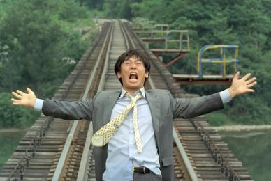 man in suit running across wooden bridge with arms outstretched and mouth open