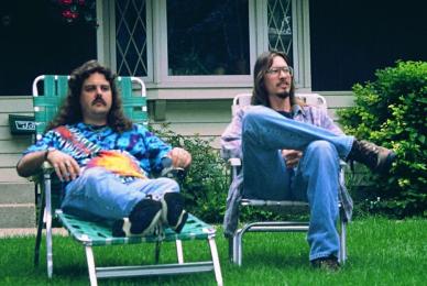 two men in jeans and tie dye shirts sitting in lawn chairs in grass yard